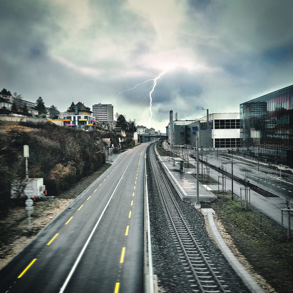 Route vide avec marquages jaunes à gauche et voie ferrée à droite, sous un ciel nuageux.