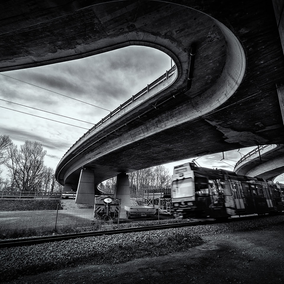 Pont autoroutier sinueux au-dessus d'une voie ferrée avec un train, arbres en arrière-plan, ciel nuageux.