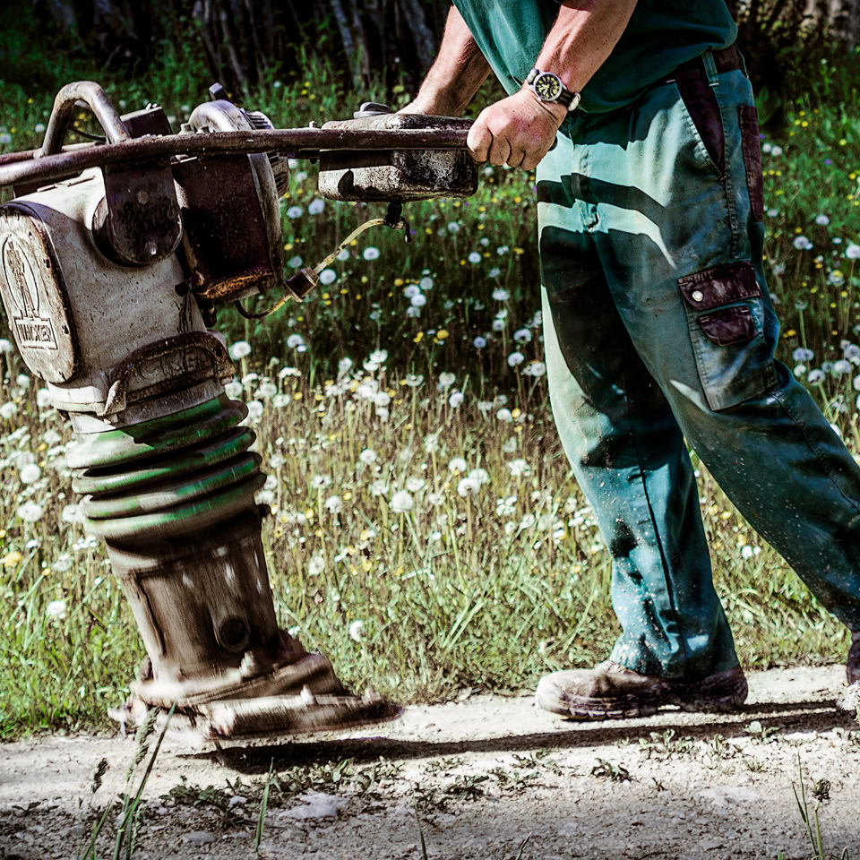 Personne utilisant une machine à compacter sur un chemin de terre, portant des vêtements de travail verts.