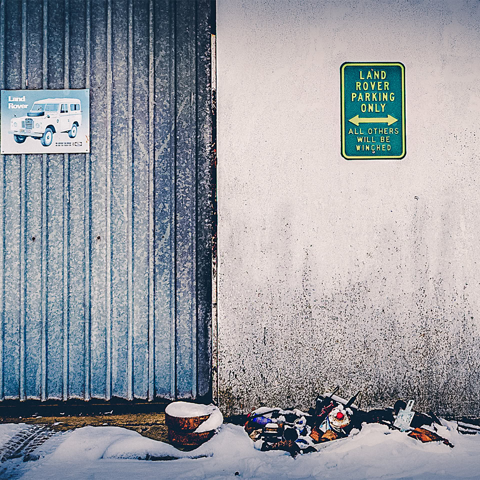 Mur extérieur avec une porte métallique bleue, deux affiches, un panneau vert, et des fleurs déposées au sol.
