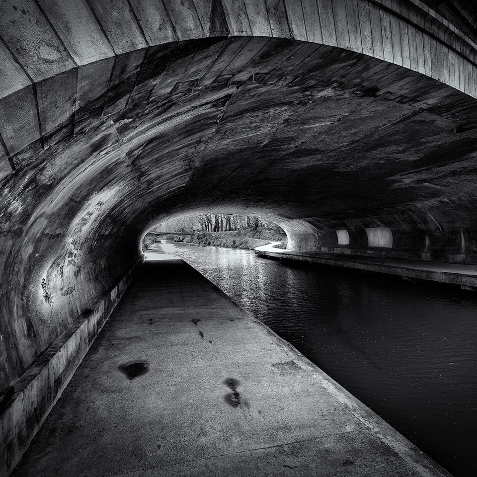 Vue en noir et blanc d'un tunnel avec un chemin piétonnier à gauche et un canal d'eau à droite.