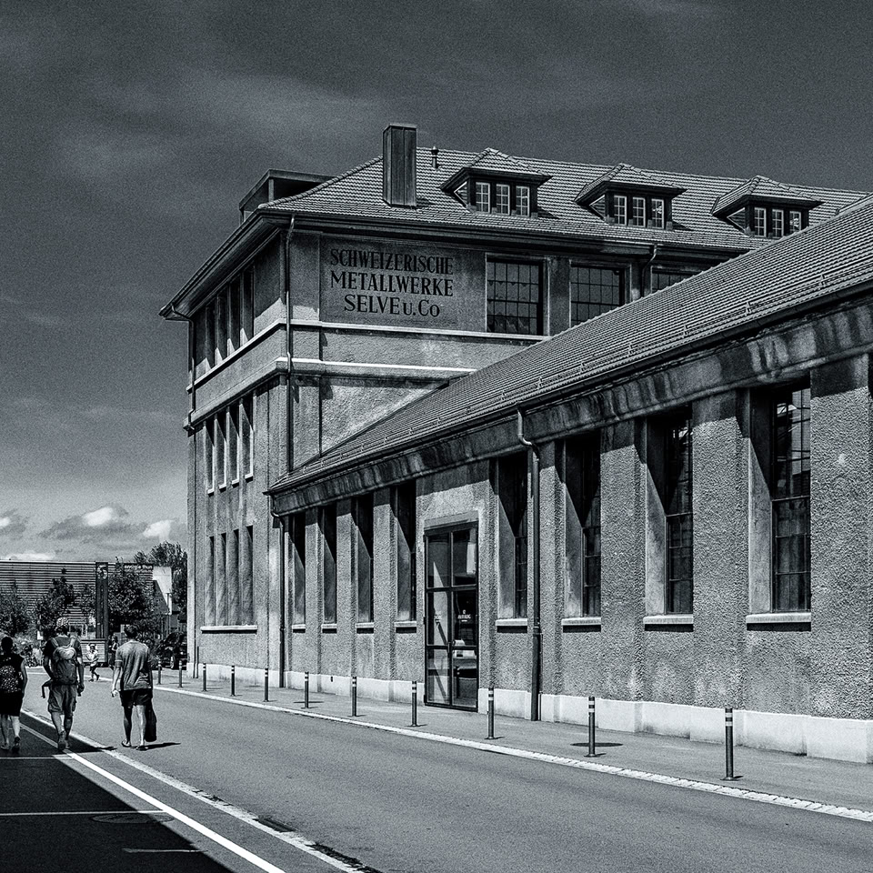 Rue avec un grand bâtiment en briques à droite, quelques personnes marchant sur le trottoir, ciel nuageux.