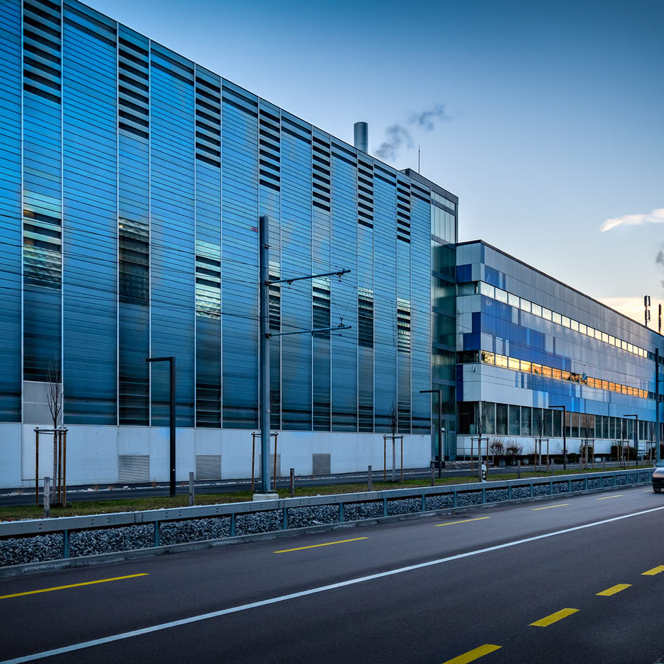 Bâtiment moderne en verre avec une route au premier plan et une voiture en mouvement.