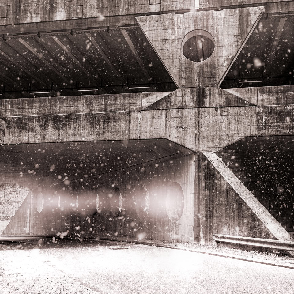 Snow falling under a concrete bridge with a road beneath, captured in black and white.