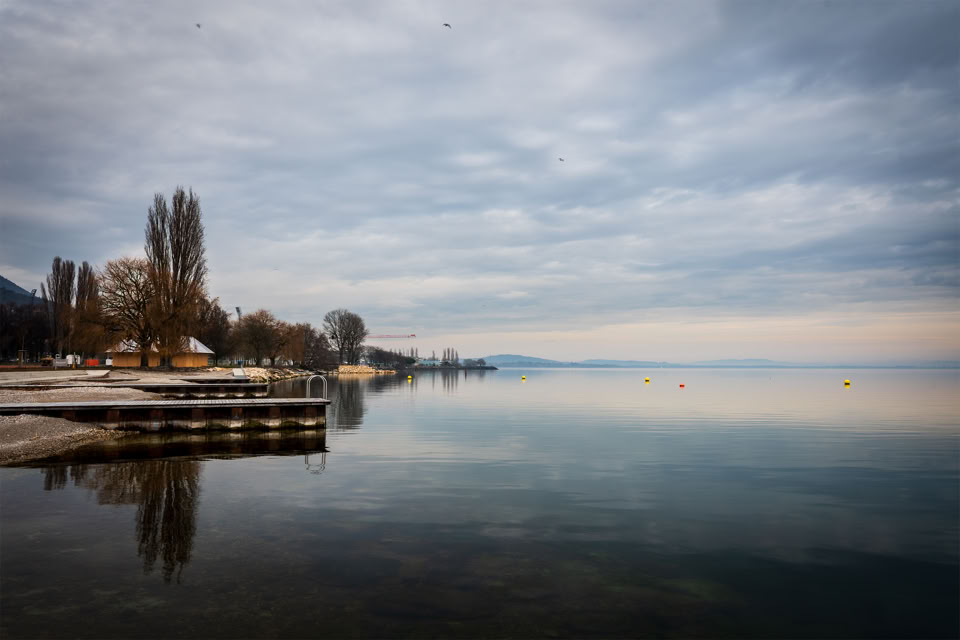 A calm lake with a wooden dock on the left, surrounded by bare trees and a cloudy sky.