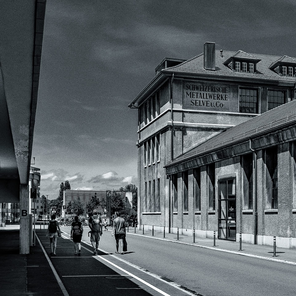 A black and white image of a street with a large brick building on the right, featuring arched windows. Several people are walking along the sidewalk. The sky is partly cloudy.