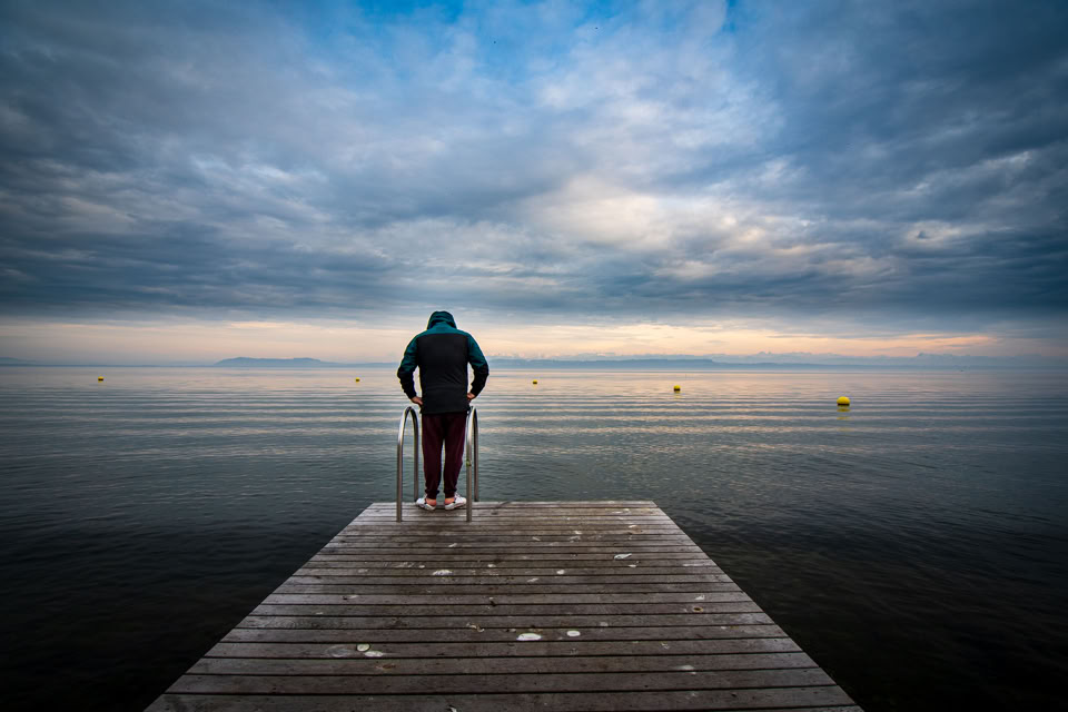 Une personne debout au bout d'un quai en bois, regardant vers un lac sous un ciel nuageux.
