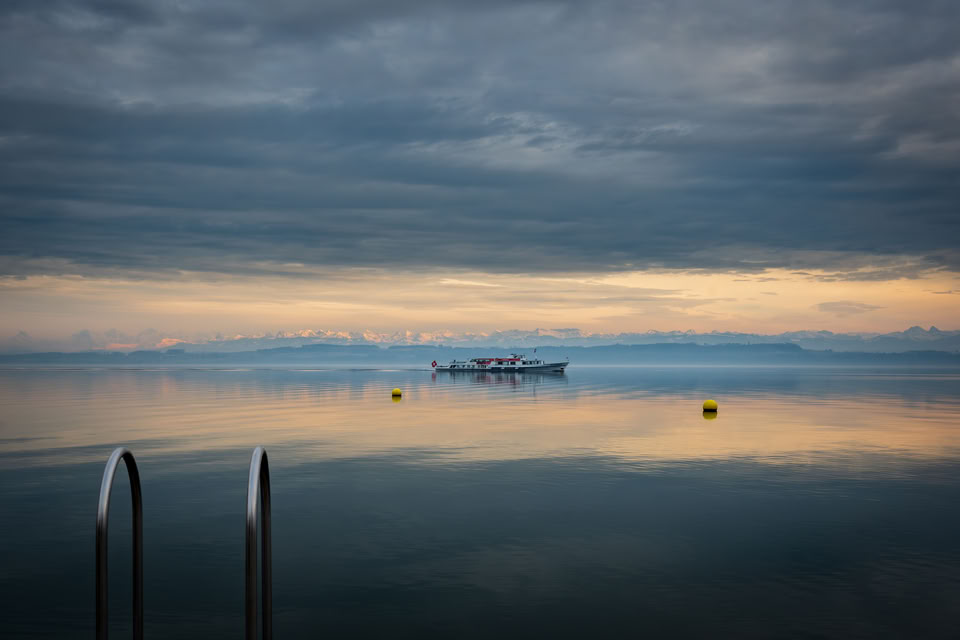 A calm body of water with a distant boat, two yellow buoys, and a cloudy sky. A metal ladder is visible in the foreground.