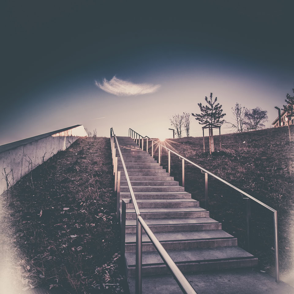 A concrete staircase with metal railings leading up a grassy slope, with a single cloud in the sky and trees in the background.