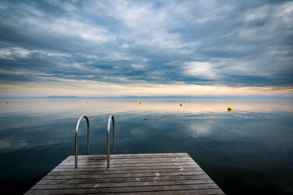 Wooden dock with metal ladder leading into a calm body of water under a cloudy sky.