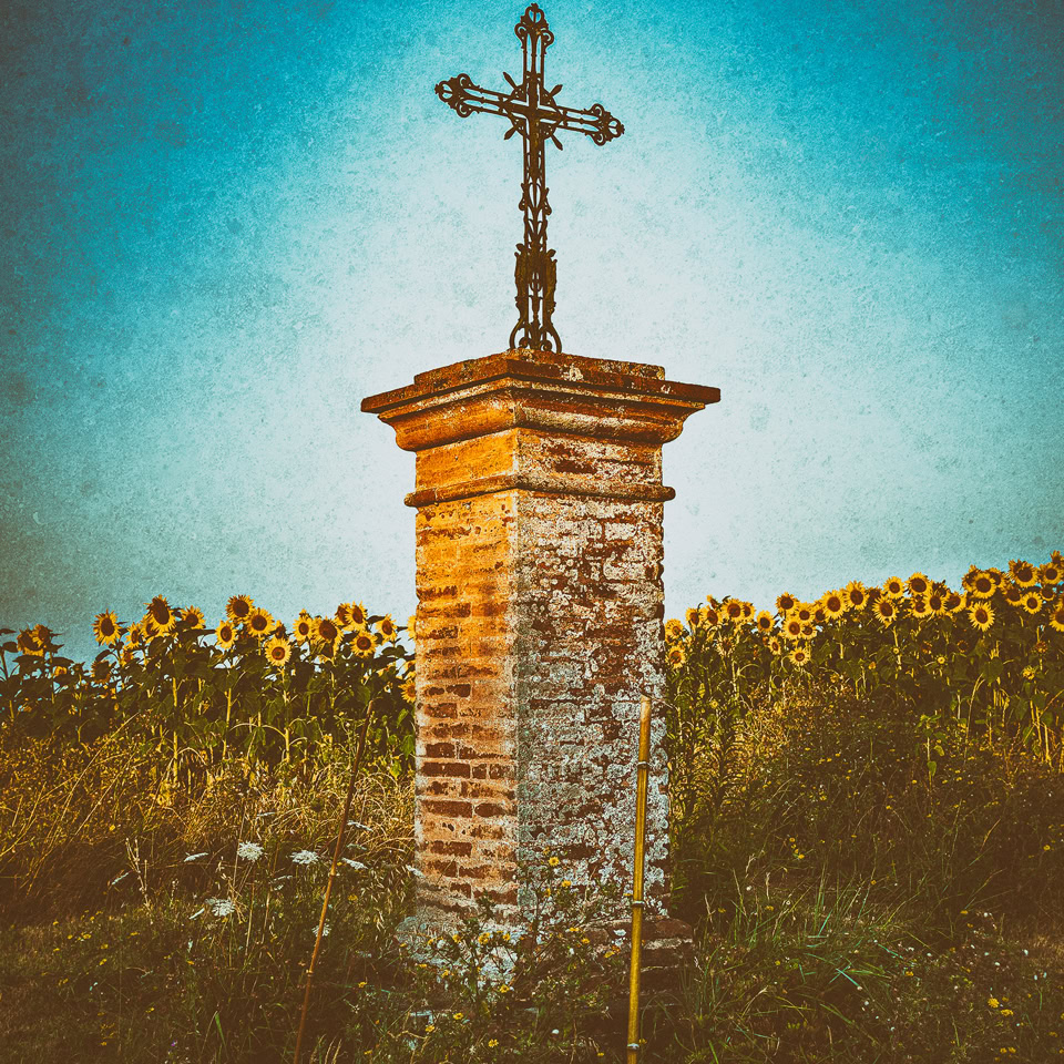 Croix en métal sur un pilier en pierre, entourée de tournesols sous un ciel bleu.