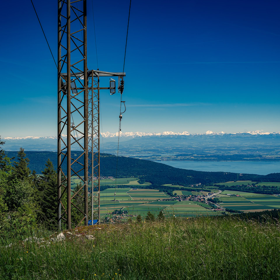 Paysage avec un pylône électrique, des arbres, une prairie verte, et une vue sur un lac et des montagnes à l'horizon sous un ciel bleu.