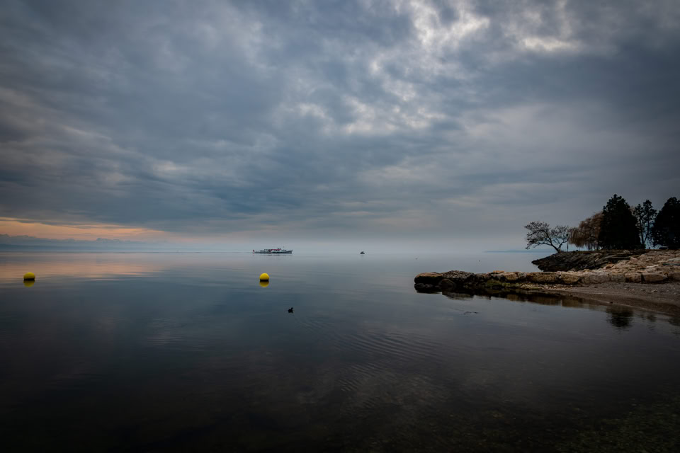 Paysage lacustre avec ciel nuageux, eau calme, bouées jaunes, et rive avec arbres à droite.